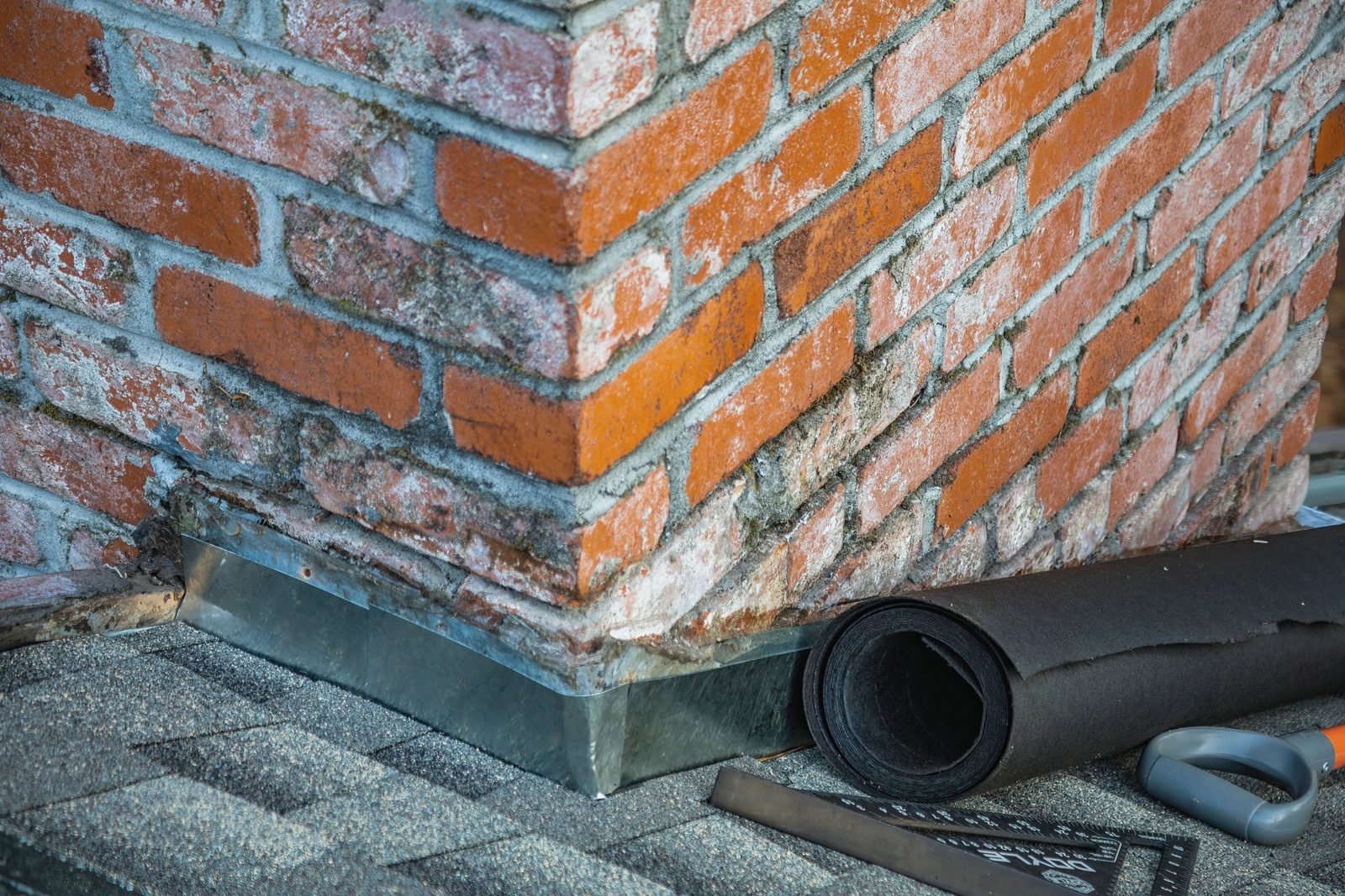 New metal flashing at the base of a brick chimney, with roofing felt and a square nearby
